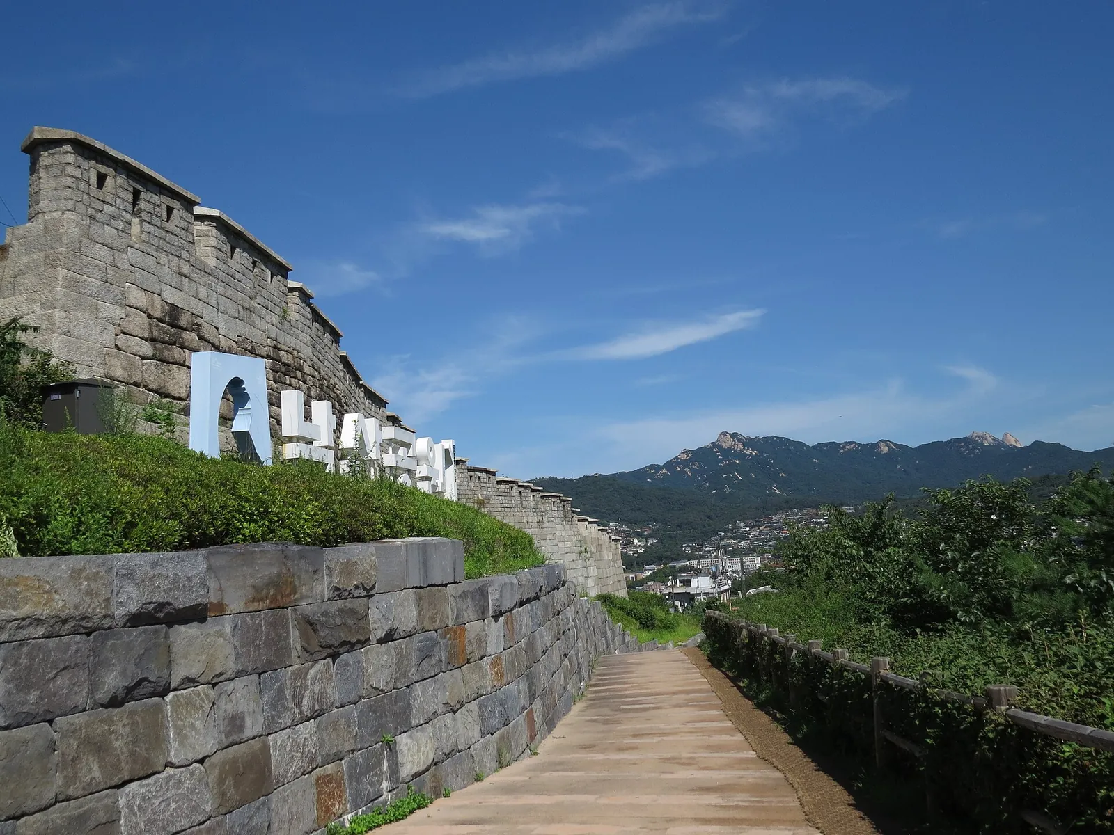 A paved path beside the stone Seoul City Wall on Naksan, with Naksan Park lettering on the wall and mountains on the horizon under a blue sky