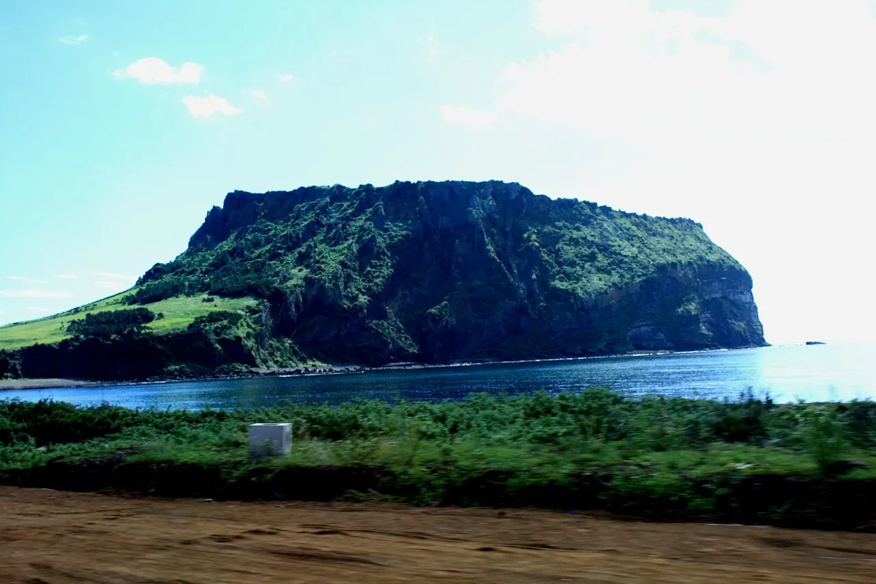 Seongsan Ilchulbong, a green-topped tuff cone with steep cliffs, rising from the sea under a blue sky on Jeju