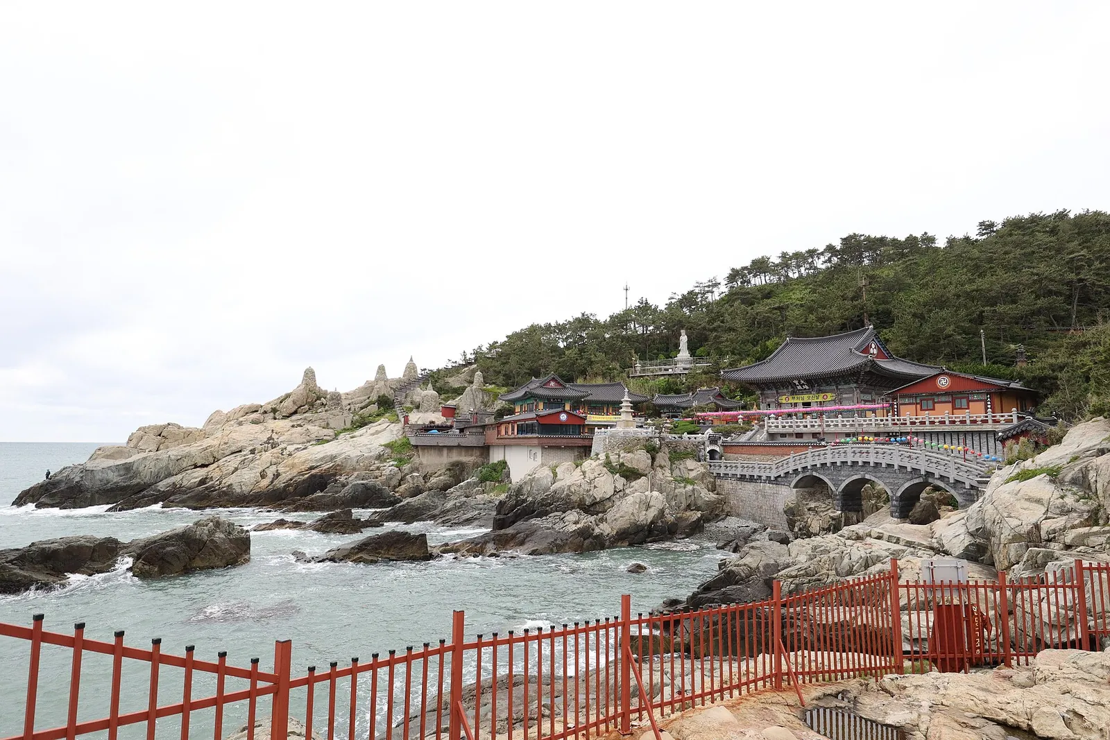 Haedong Yonggungsa temple buildings on the rocky shoreline beside the sea in Busan, seen past a red railing