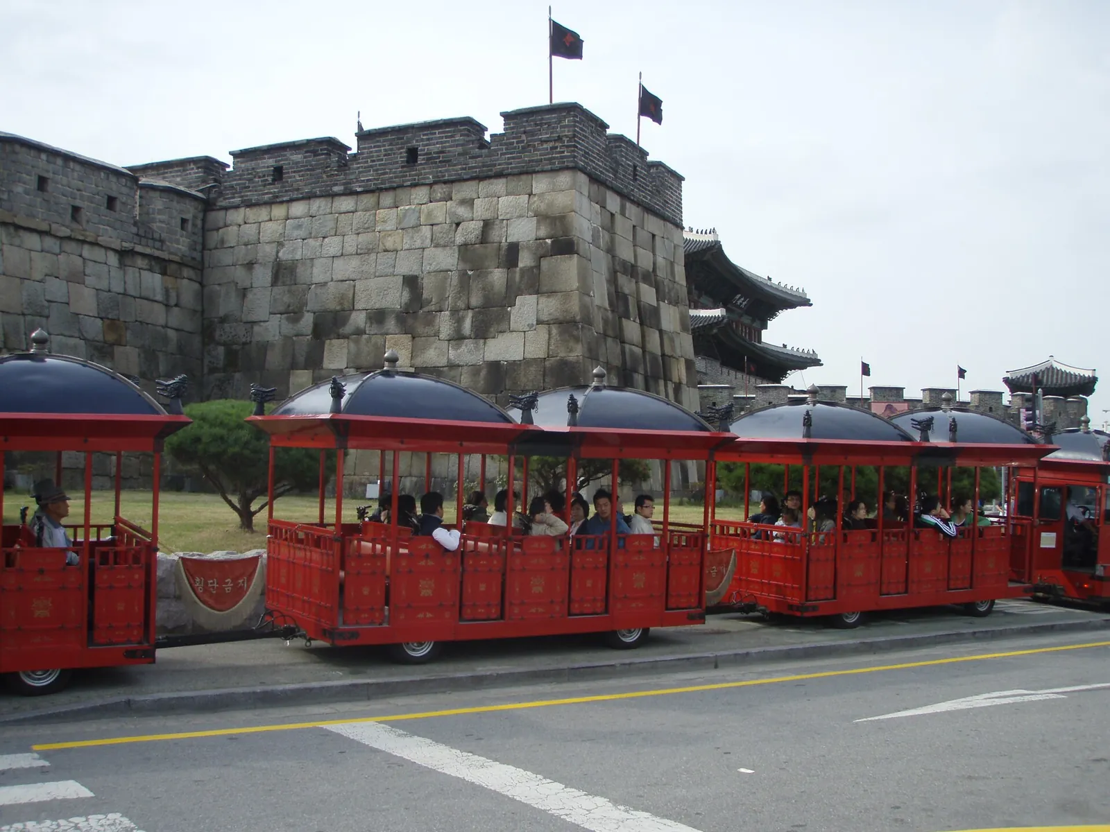 Røde, åbne turistvogne på en vej ved siden af en stenbastion på Hwaseong-fæstningen i Suwon