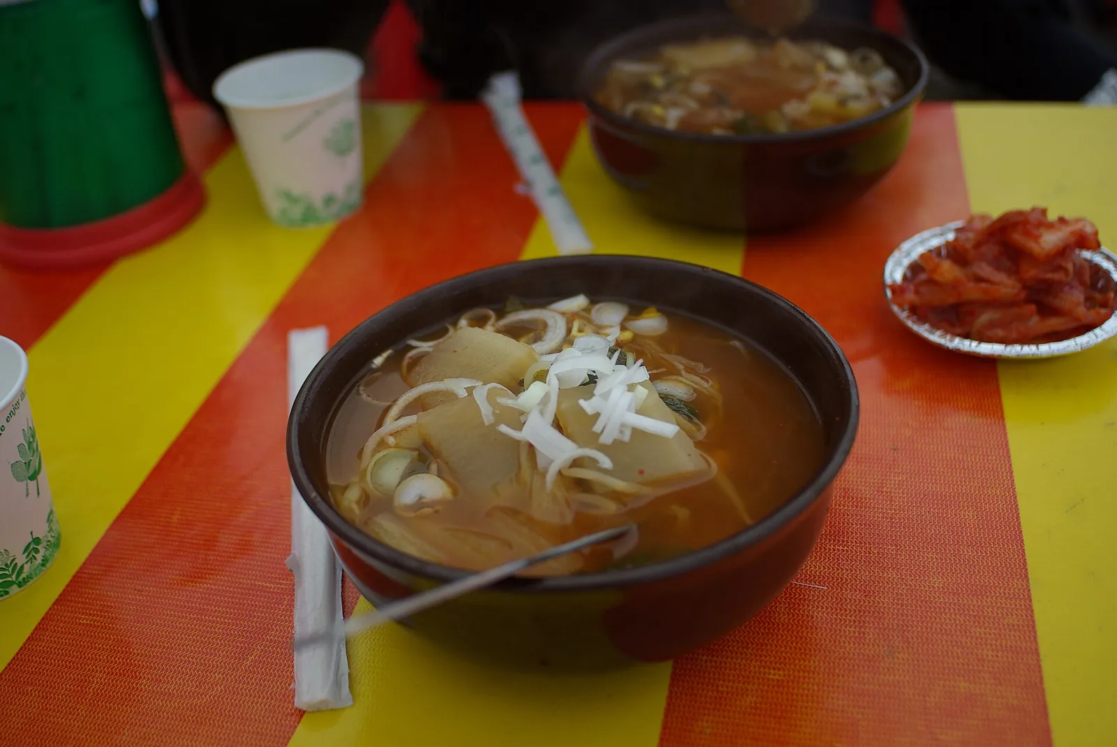 A dark bowl of gukbap, Korean rice soup, with sliced radish and spring onion in broth, and a dish of kimchi on the side
