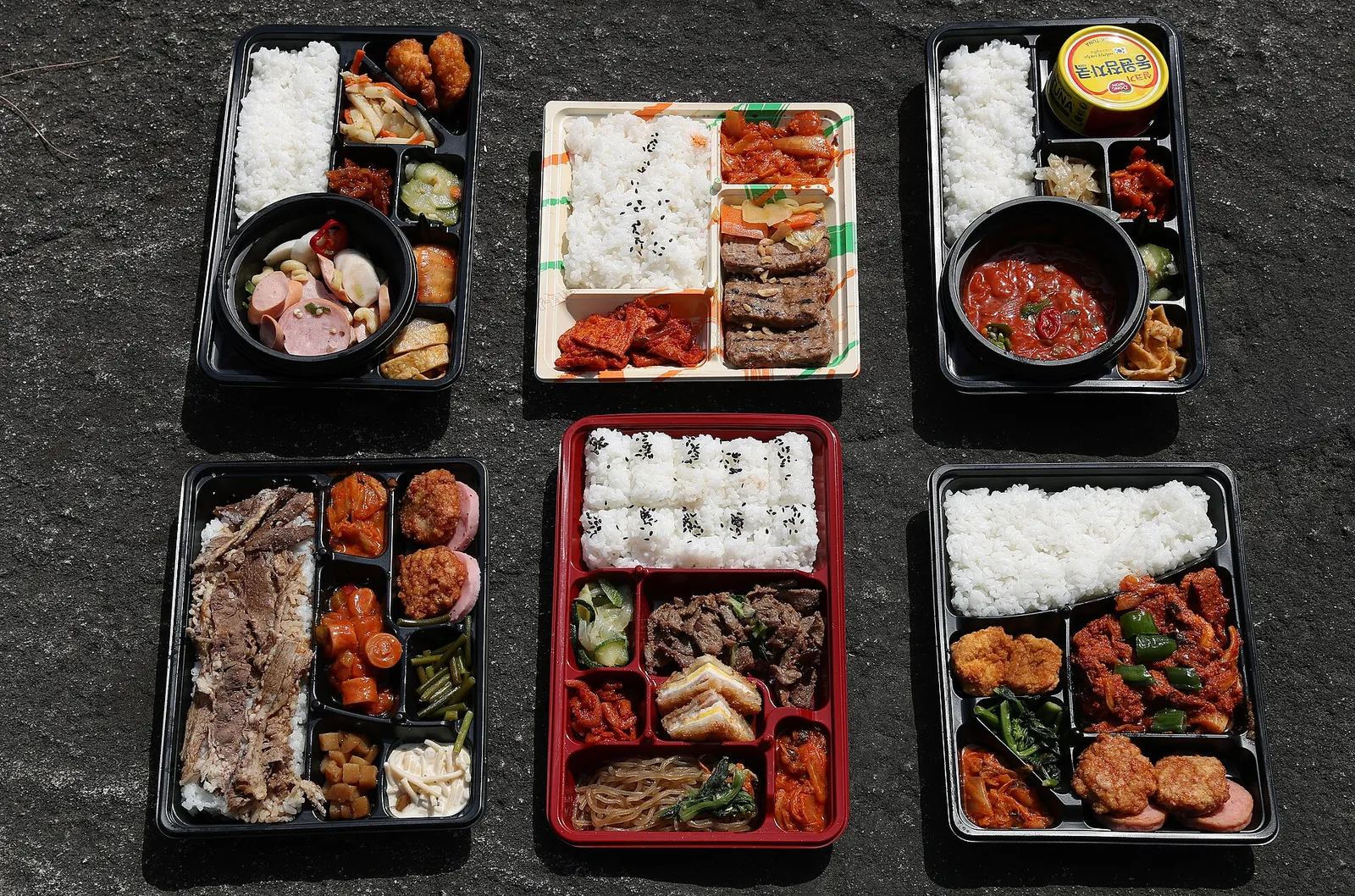 Six Korean lunch boxes from a convenience store, with rice, grilled meat, kimchi and side dishes in black trays
