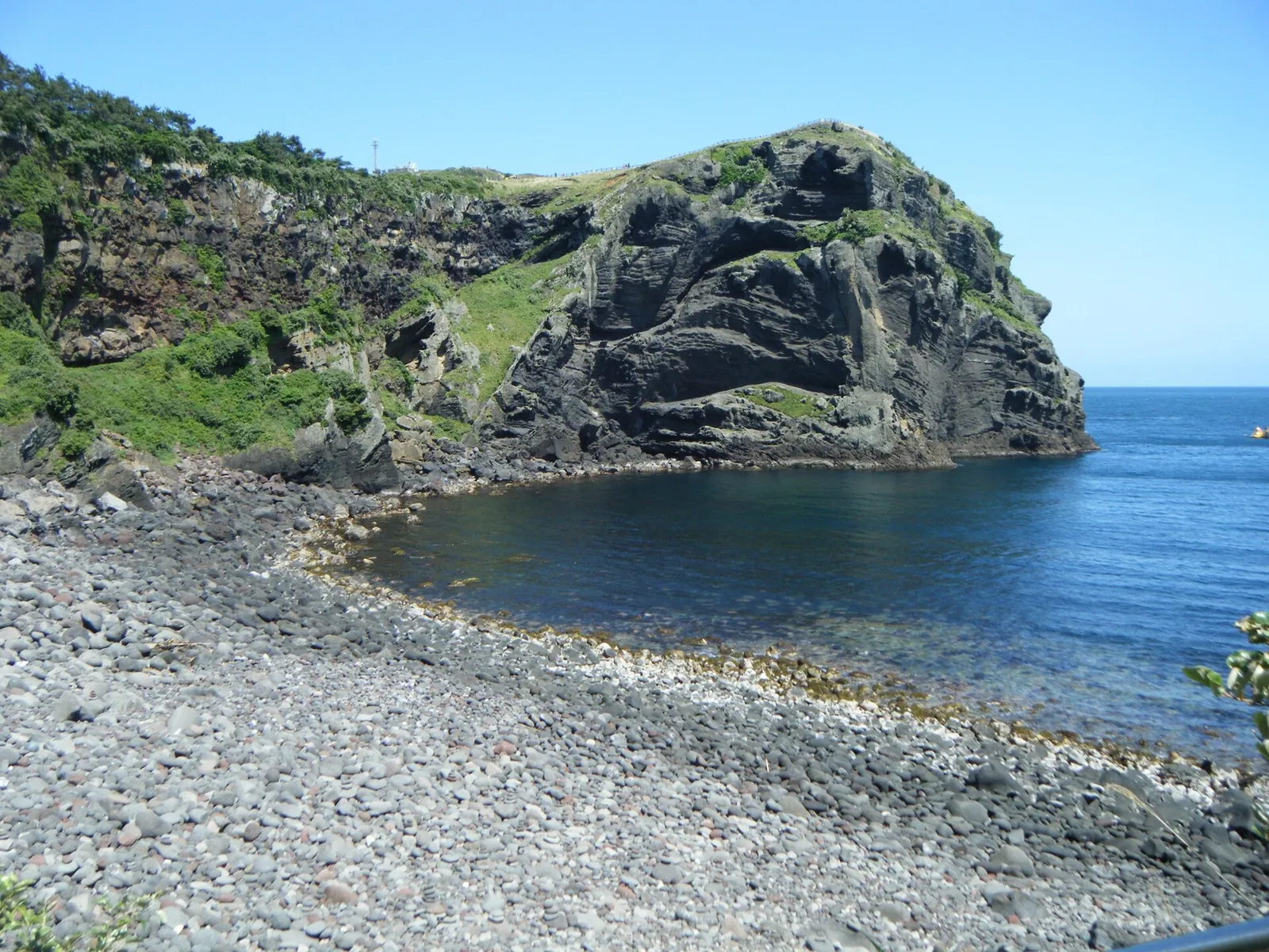 Dark volcanic cliffs and green slopes above a grey pebble shore and calm blue water on Udo