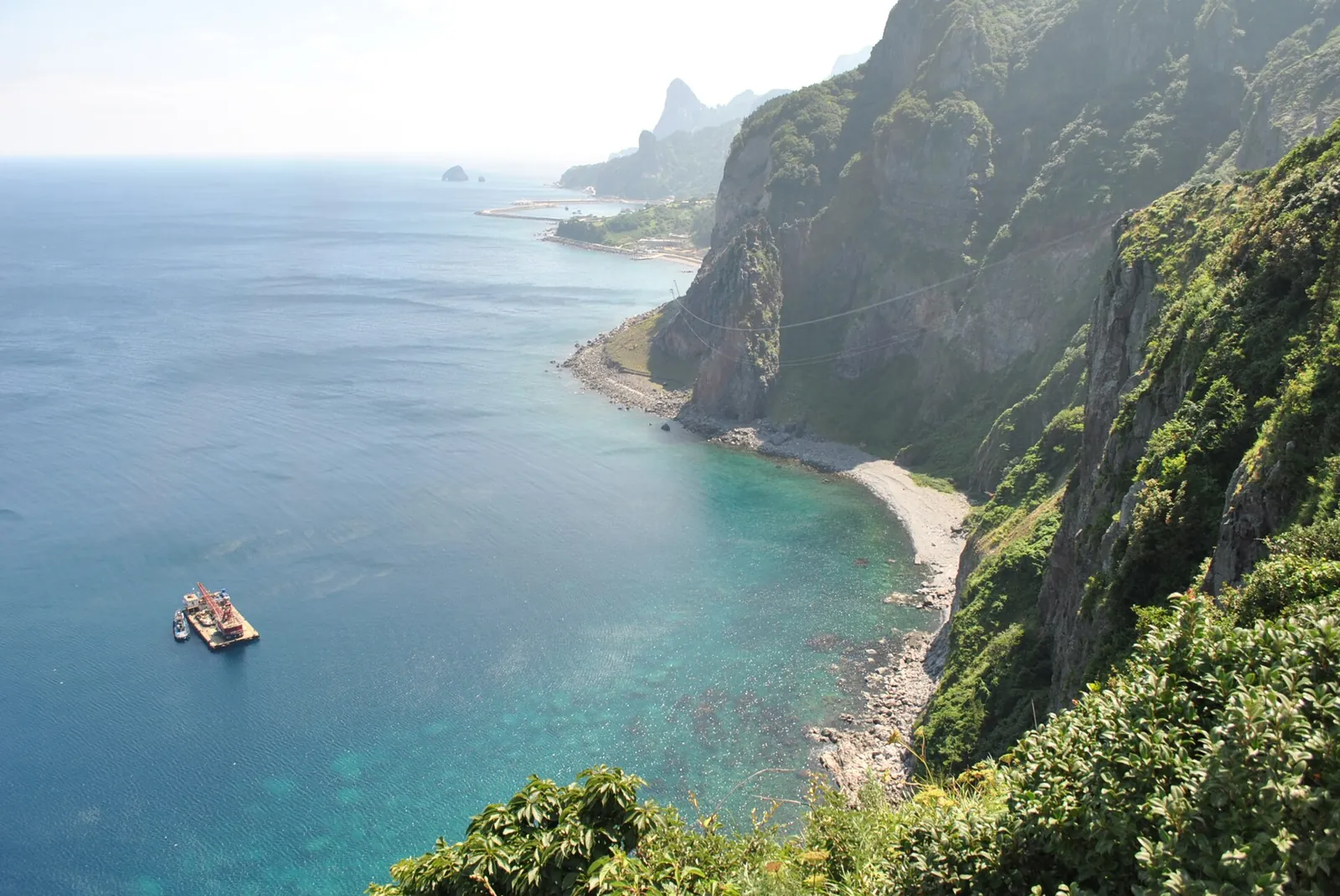 Green volcanic cliffs dropping into turquoise water near Taeha on Ulleungdo, with a pebble shore and a small boat below