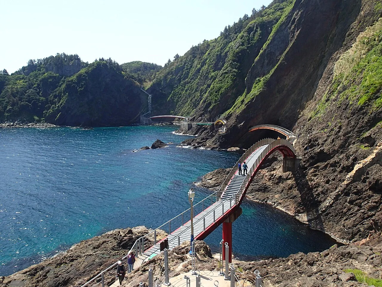 An arched footbridge on a coastal walking path between dark volcanic cliffs on Ulleungdo