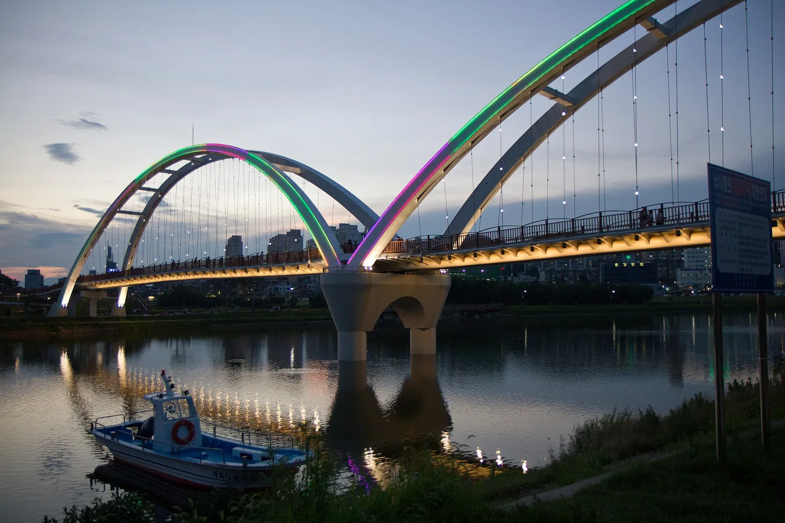 The arched Simnidaebat Bridge over the Taehwagang in Ulsan, lit in colour at dusk, with a small boat moored below