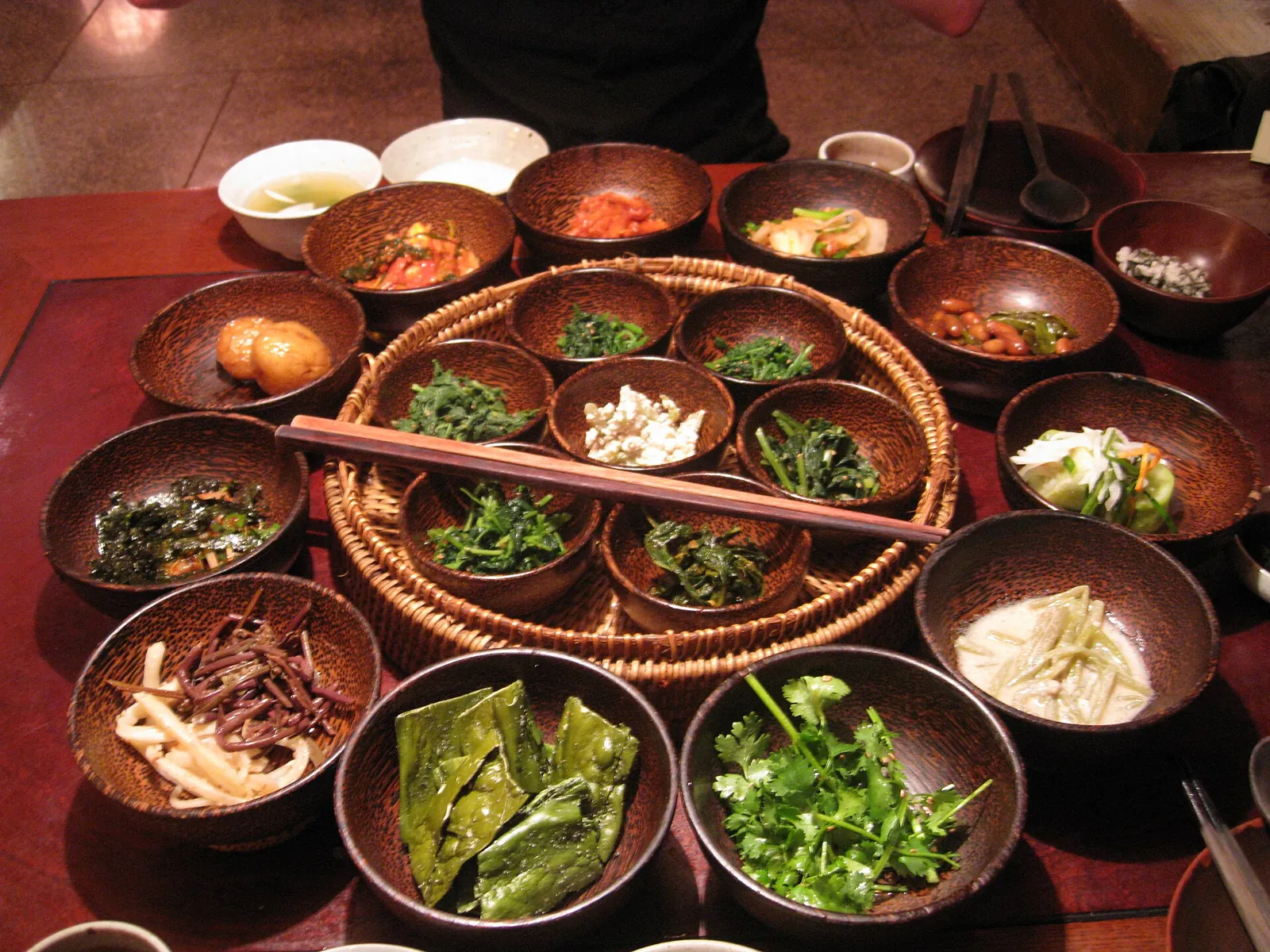Many small bowls of seasoned greens, roots and vegetables arranged around a round basket tray on a restaurant table in Insa-dong, Seoul