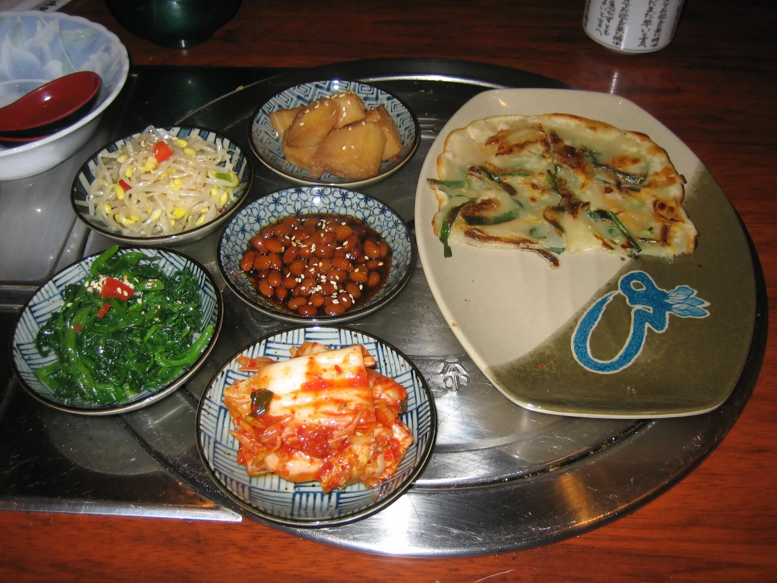 A green-onion pancake on a metal tray with small side dishes of kimchi, bean sprouts, seasoned greens and braised beans