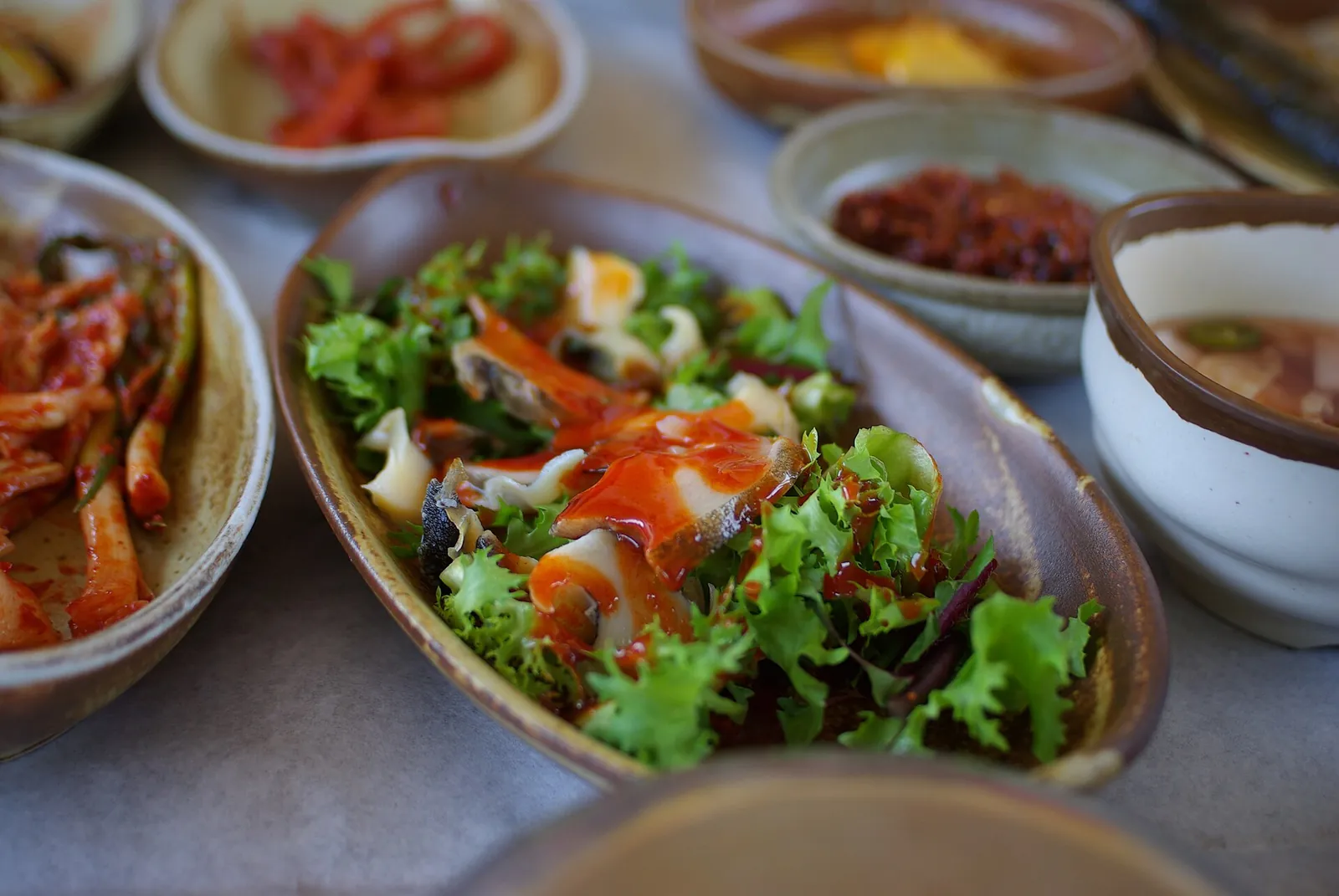 A dish of sliced raw abalone with salad leaves and red chili sauce, surrounded by side dishes