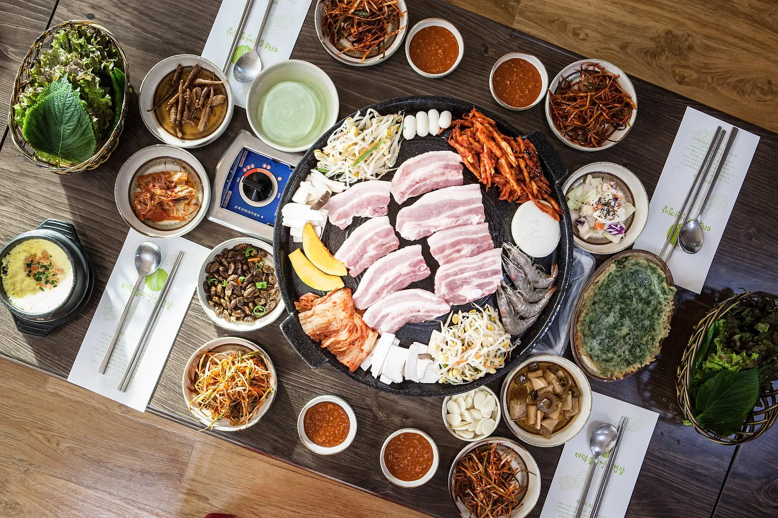 Table set for samgyeopsal with pork belly on a round grill surrounded by side dishes and lettuce