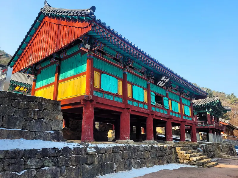 The red-and-green painted Bogwangru Pavilion of Guryongsa Temple under a blue sky, with snow on the steps