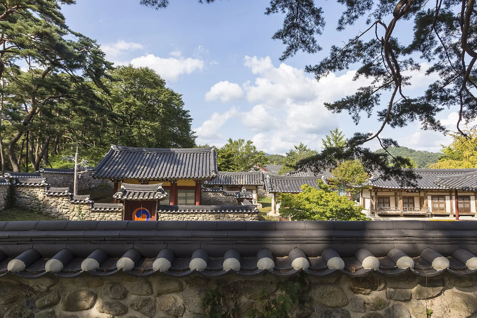 Tiled-roof buildings of Sosuseowon Confucian Academy behind a low stone wall under pine trees
