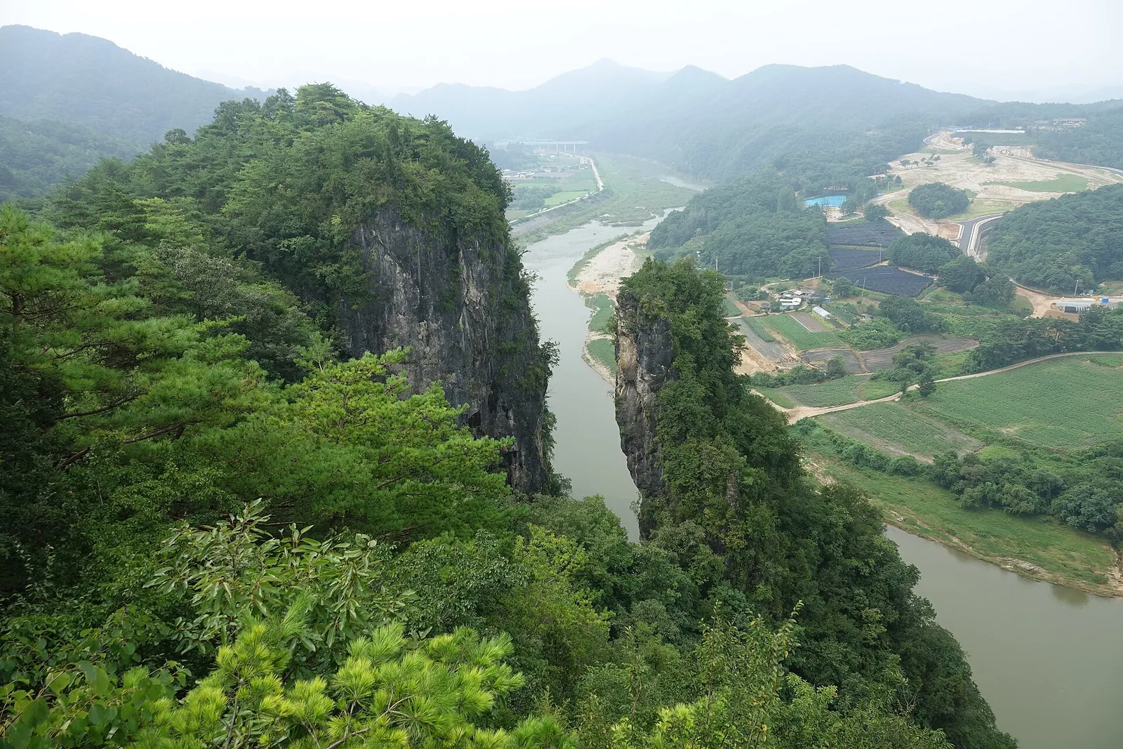 A tall rock pillar split from a forested cliff above a winding river and farmland at Seondol, Yeongwol
