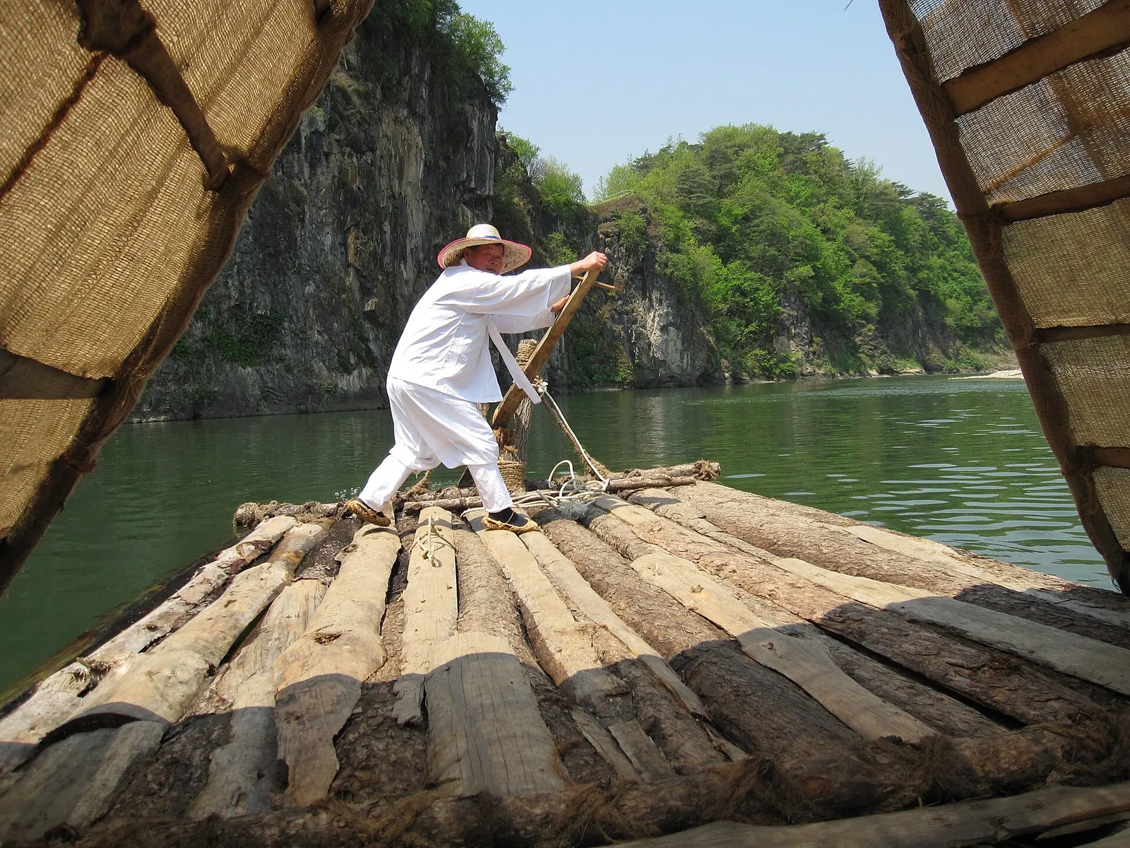 A boatman in white traditional clothes and a straw hat poles a wooden raft on a green river below a cliff in Hanbando-myeon
