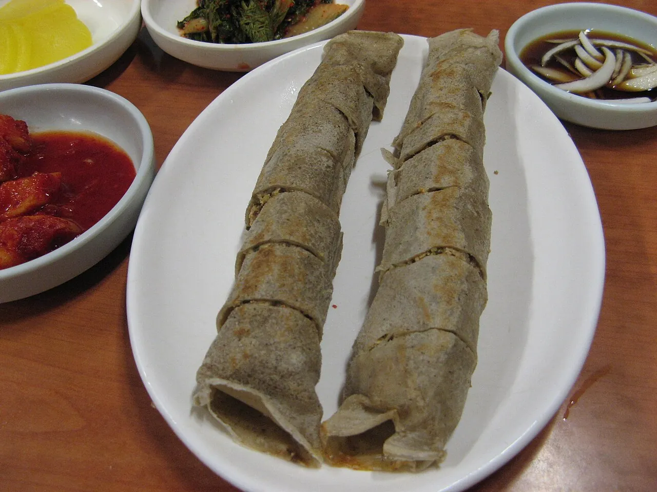Two rolled grey buckwheat crepes on a white plate, with side dishes of kimchi and dipping sauce