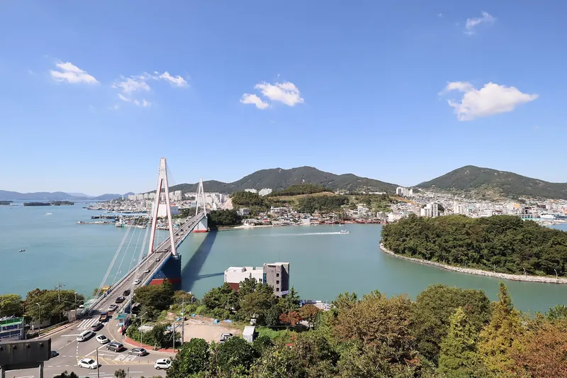 Dolsan Bridge crossing the blue harbour channel to Yeosu's hillside city under a bright sky, seen from Dolsan Park