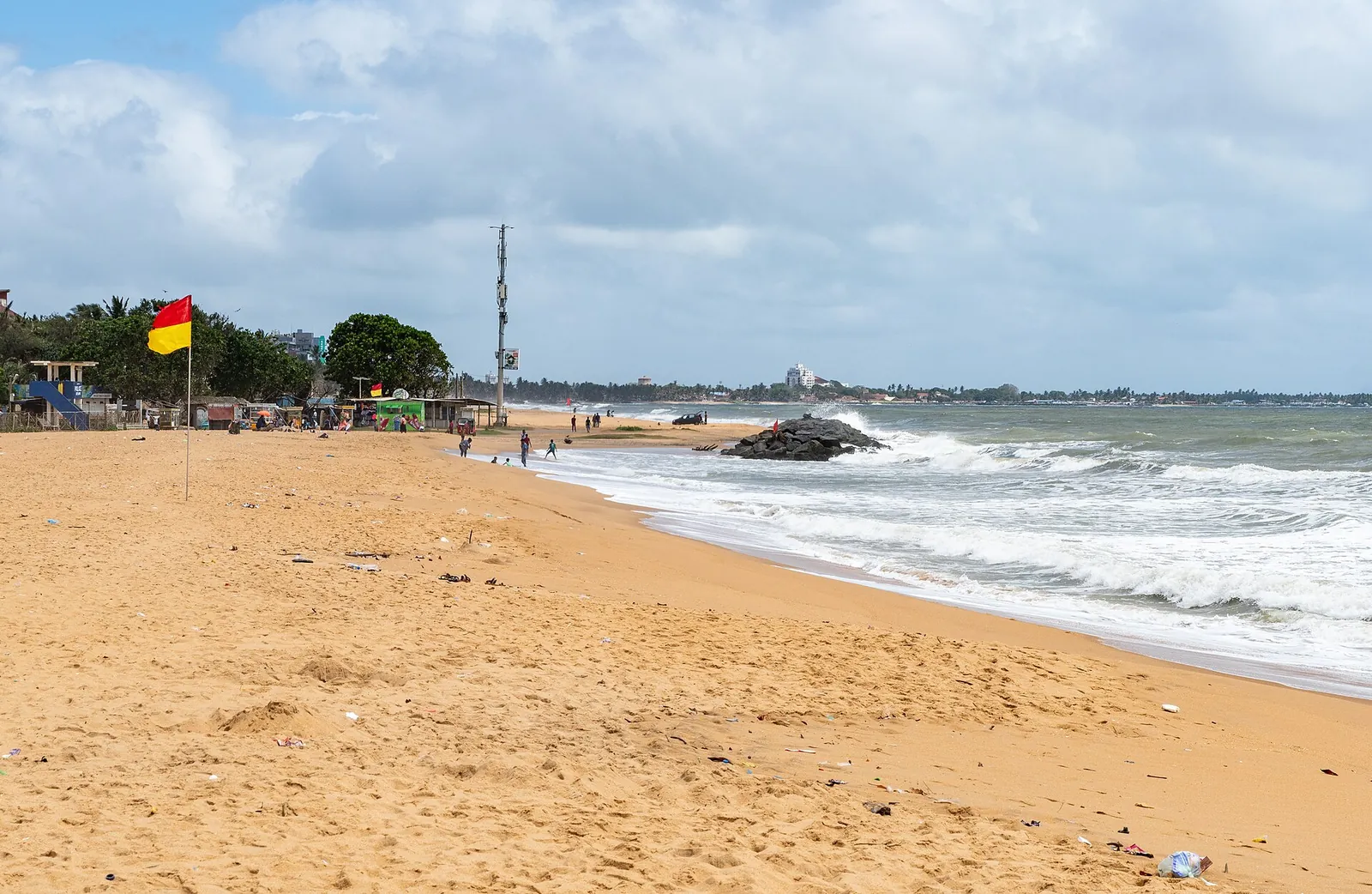 Una amplia playa de arena en Negombo con oleaje y una bandera roja y amarilla