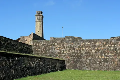 Der Uhrturm von Galle Fort ragt über eine grasbewachsene Festungsmauer unter blauem Himmel