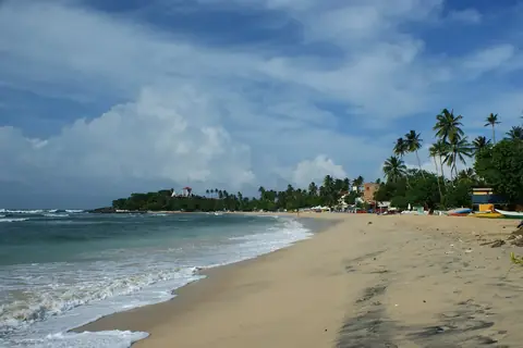 Ein Sandstrand in Unawatuna unter teils bewölktem Himmel, mit Palmen und einer niedrigen Landzunge am anderen Ende