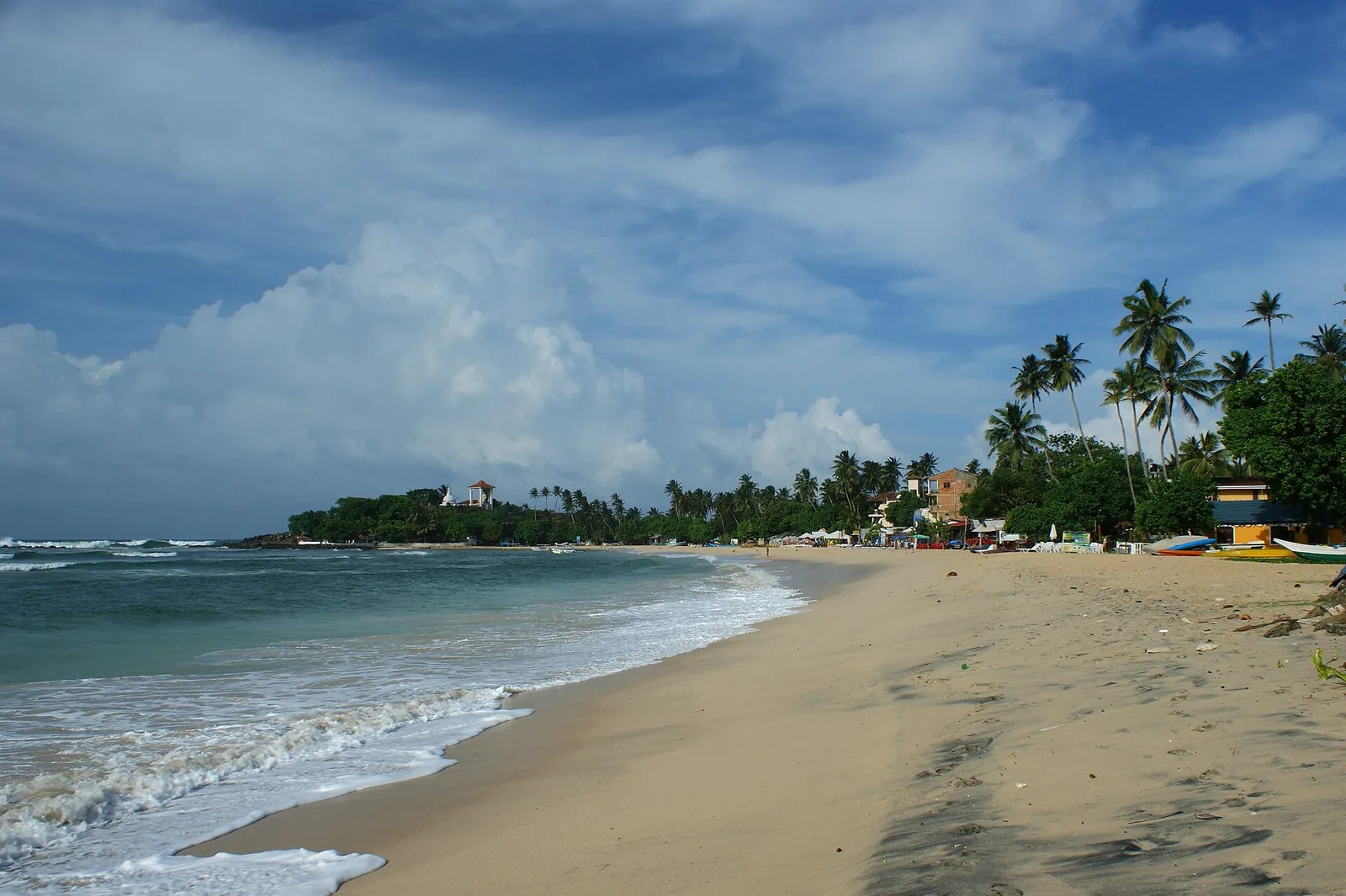 Una playa de arena en Unawatuna bajo un cielo parcialmente nublado, con palmeras y un promontorio bajo al fondo