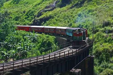 Ein roter Personenzug auf einer gebogenen Eisenbrücke über grünen Hügeln nahe dem Bahnhof Demodara