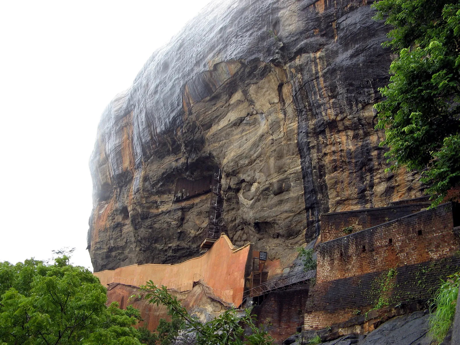 Den lodrette, regnmørke vestside af Sigiriya-klippen med en buet orange mur og en trappe ved foden, en murstensmur til højre og grønne træer rundt omkring