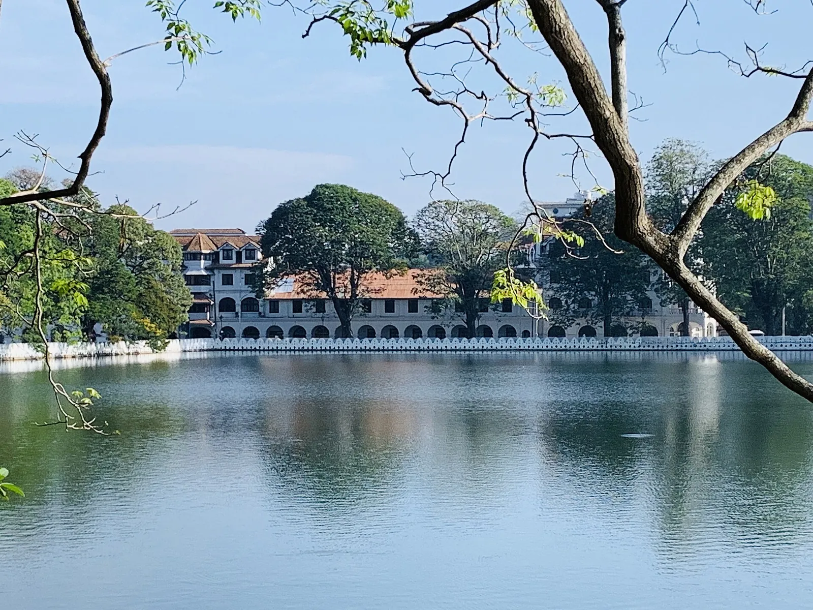 Queen's Hotel v Kandy při pohledu přes klidnou hladinu jezera Kandy se stromy v popředí
