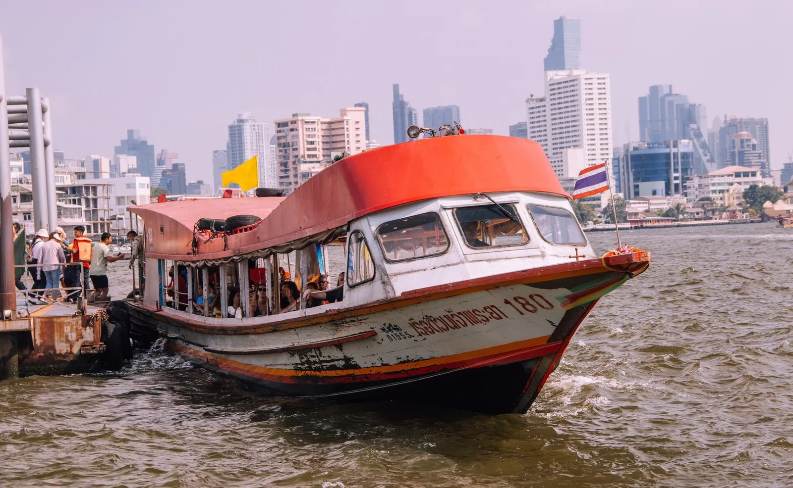 En Chao Phraya Express-båt med gult flagg ved Ratchawong-brygga, med Bangkoks høyhus i bakgrunnen