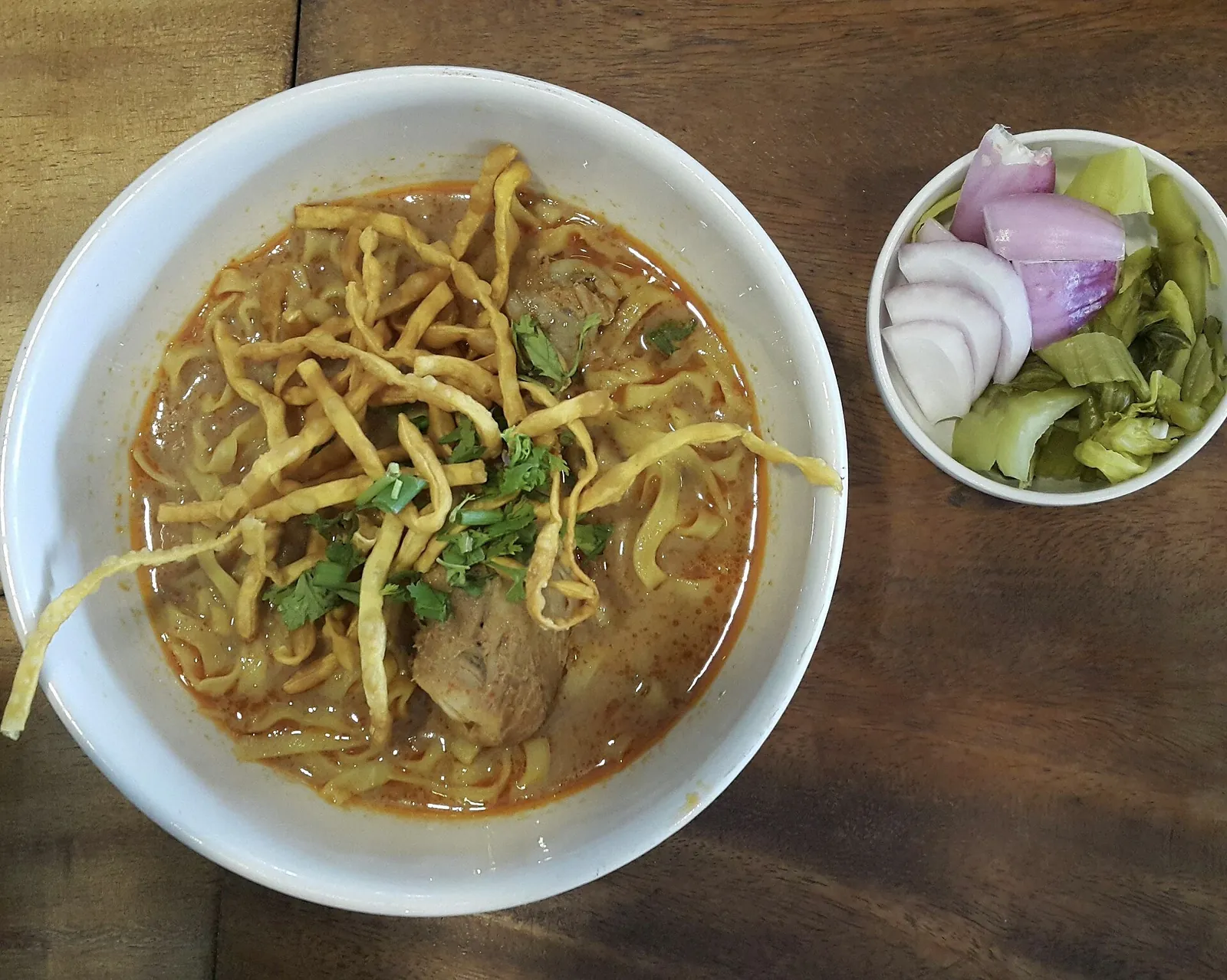 A bowl of khao soi curry noodles topped with crispy noodles beside pickled cabbage and shallots