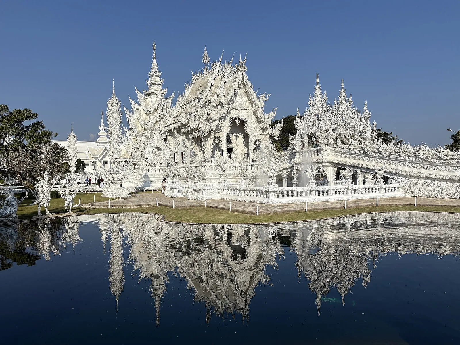 The white, mirror-decorated buildings of Wat Rong Khun reflected in a pond under a blue sky