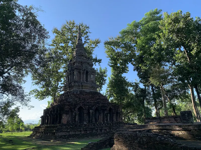 A ruined brick chedi with stucco niches under tall trees at Wat Pa Sak