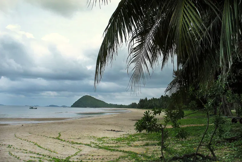 The long sweep of Thung Wua Laen beach with palms and a headland under a cloudy sky