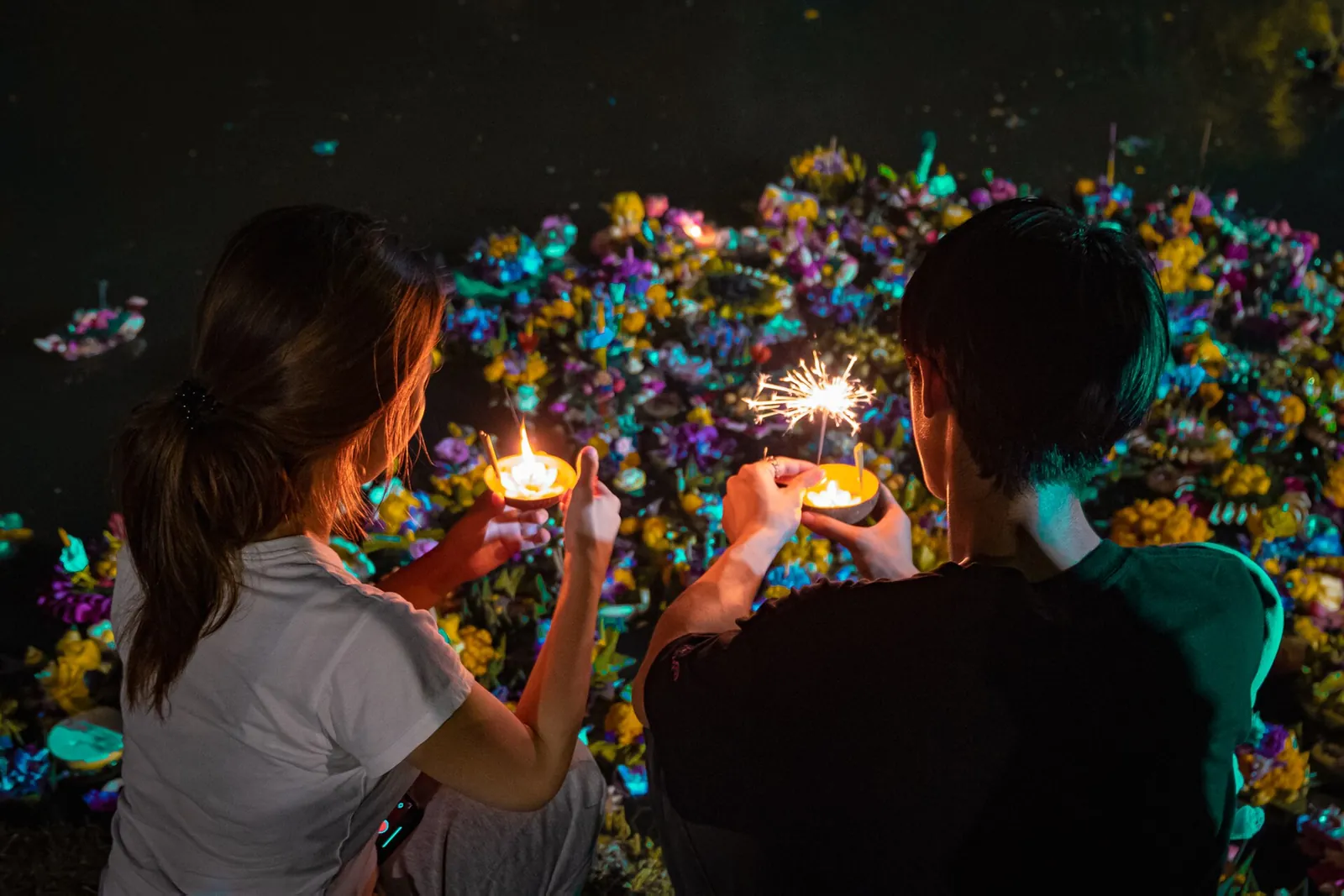 Dva lidé drží malé rozsvícené obětiny krathong u vody plné plovoucích květin v noci