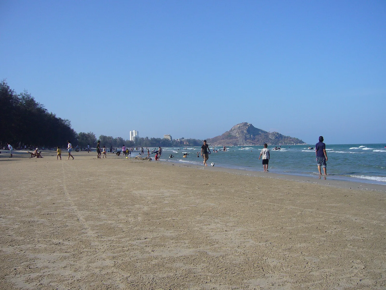 A long sandy beach with people walking at the water's edge and Khao Takiab hill in the distance