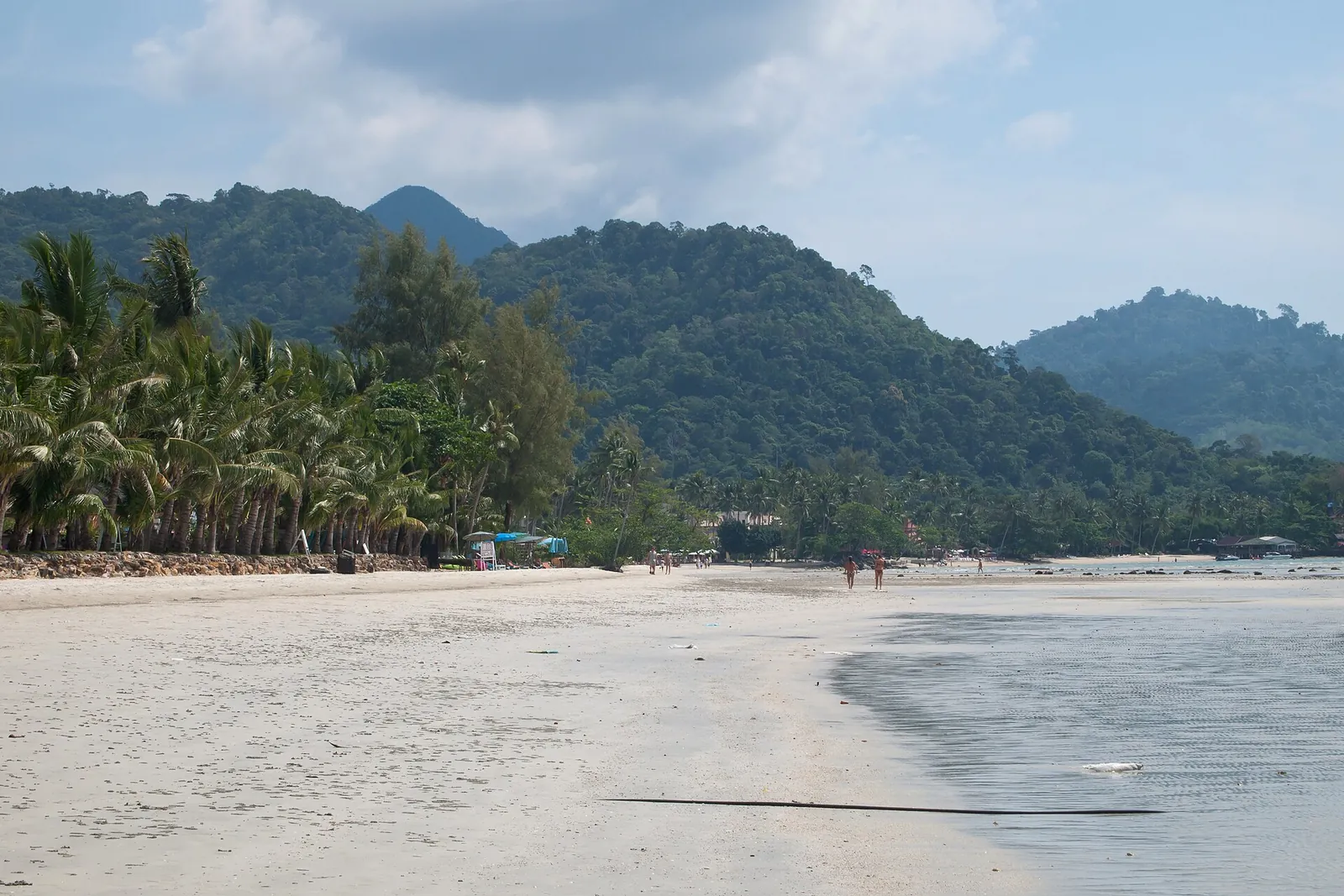 Ein breiter weißer Sandstrand mit Palmen und bewaldeten Bergen dahinter an der Westküste von Koh Chang
