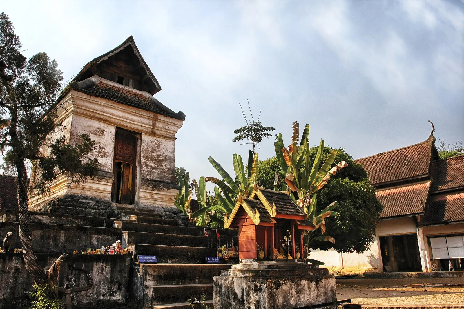 Ein alter Ziegelschrein mit Ziegeldach zwischen Bäumen auf dem Gelände des Wat Phra That Lampang Luang