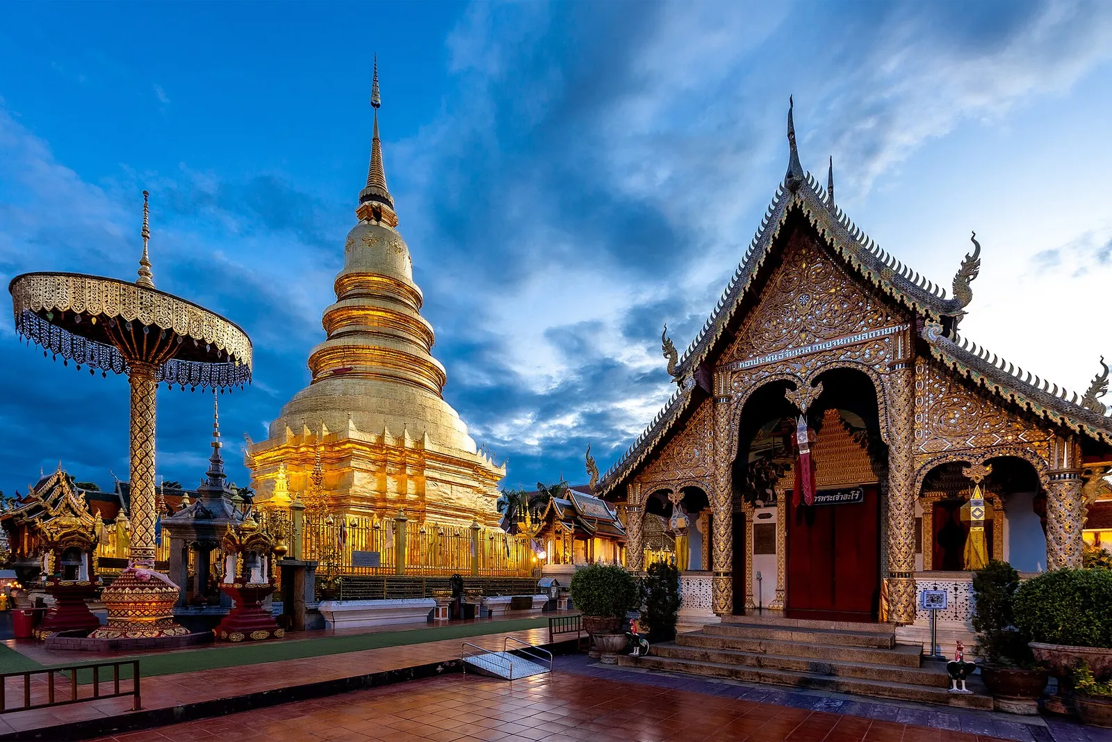 The golden main chedi of Wat Phra That Hariphunchai beside a gilded parasol and a carved hall