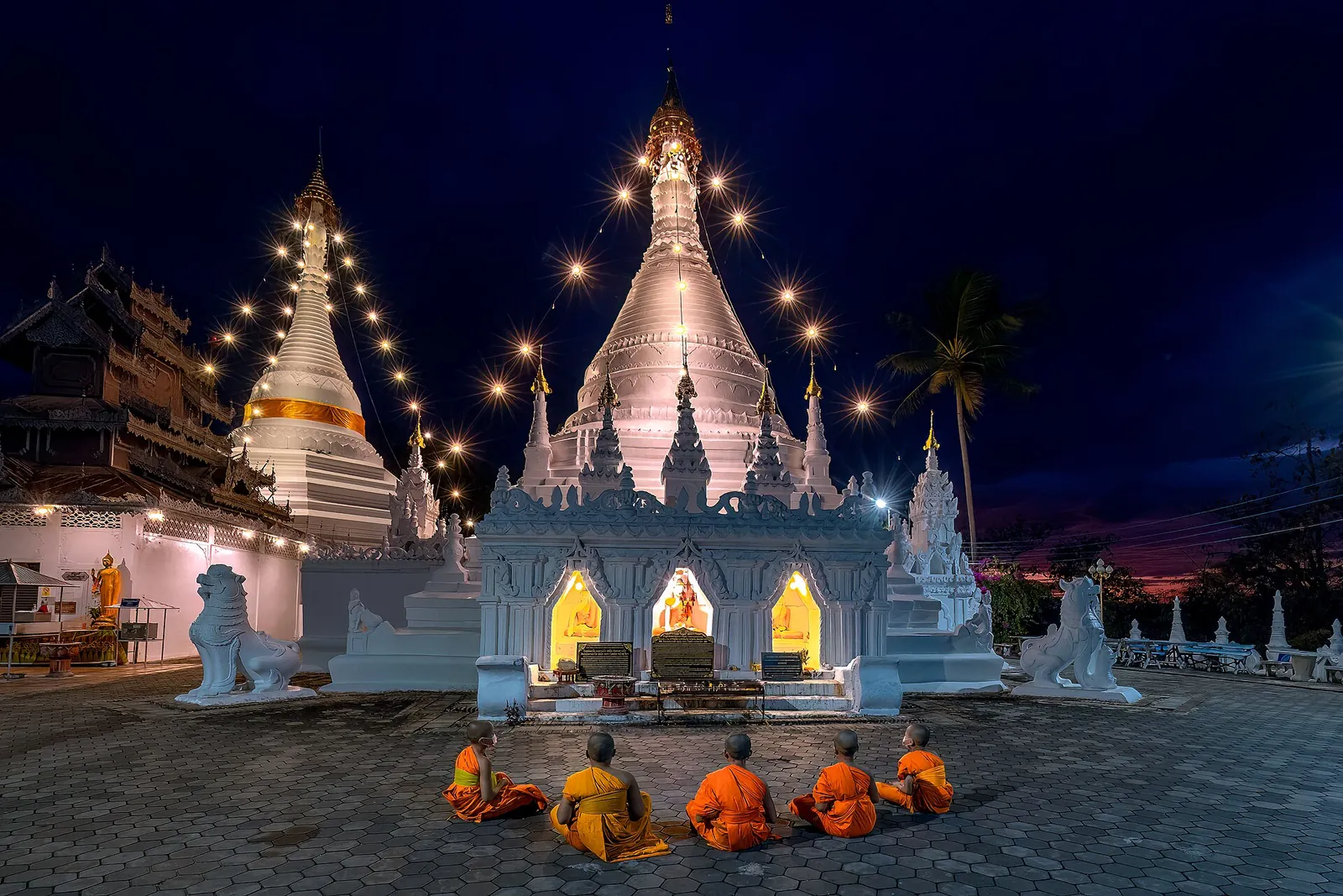 Mnisi w pomarańczowych szatach przed białymi chedi Wat Phra That Doi Kong Mu podświetlonymi o zmierzchu
