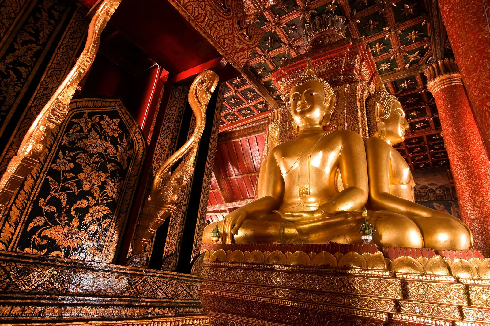 Golden Buddha images seated back to back under a carved red and gold ceiling inside Wat Phumin