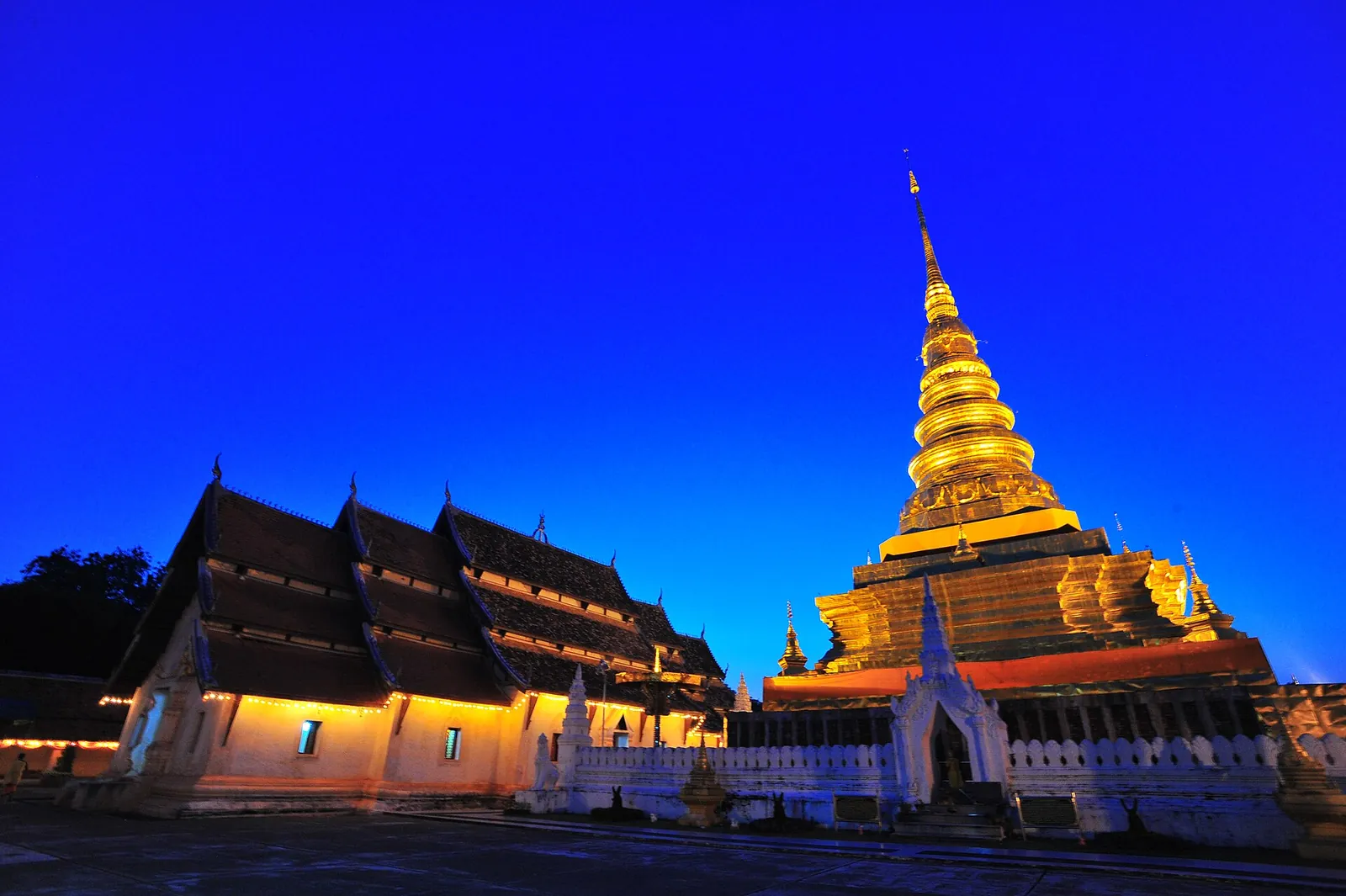 The golden chedi of Wat Phra That Chae Haeng lit at dusk beside a Lanna hall