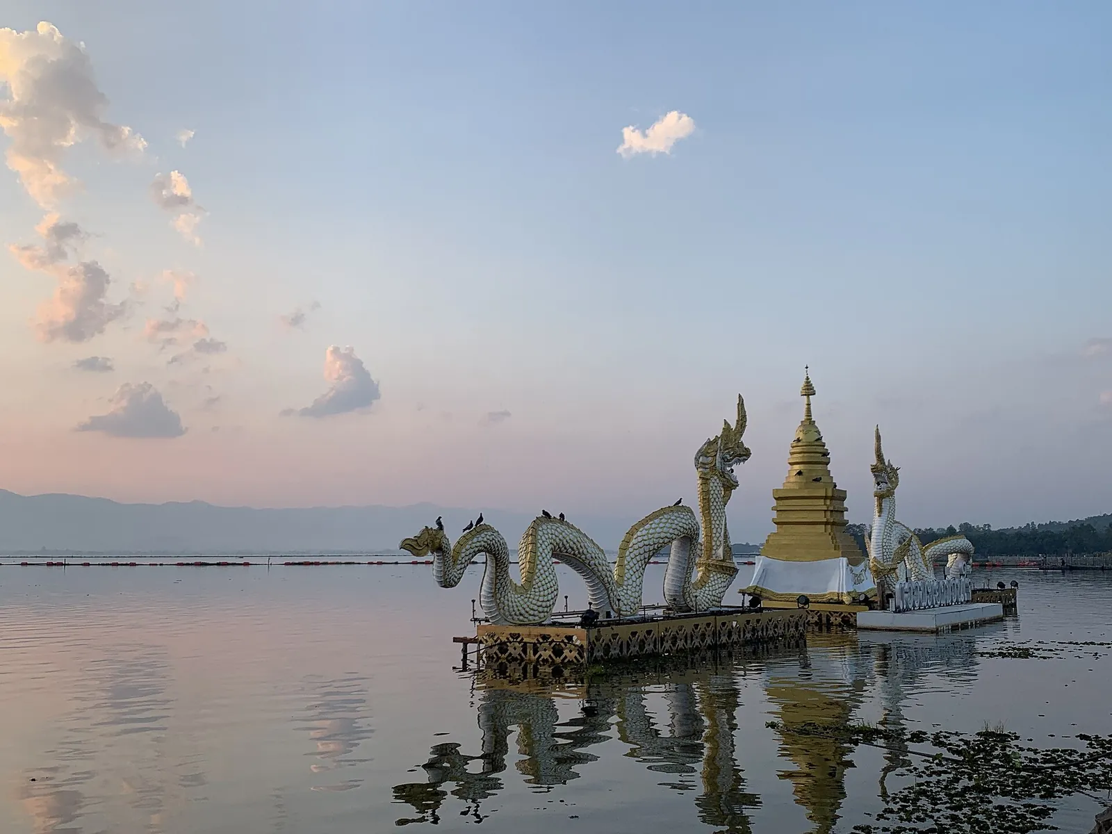 Esculturas de nagas e um pequeno chedi dourado no Kwan Phayao sob um céu cor-de-rosa ao pôr do sol