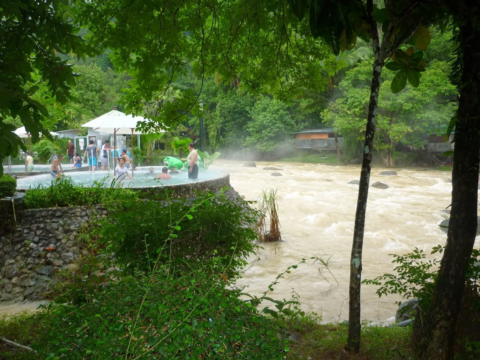 Visitors sitting by steaming hot spring pools beside a river at Raksawarin