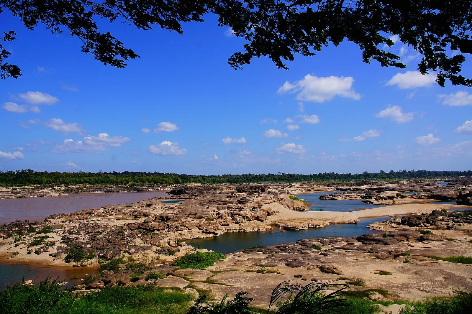 A wide riverbed of eroded sandstone and pools beside the Mekong under a blue sky