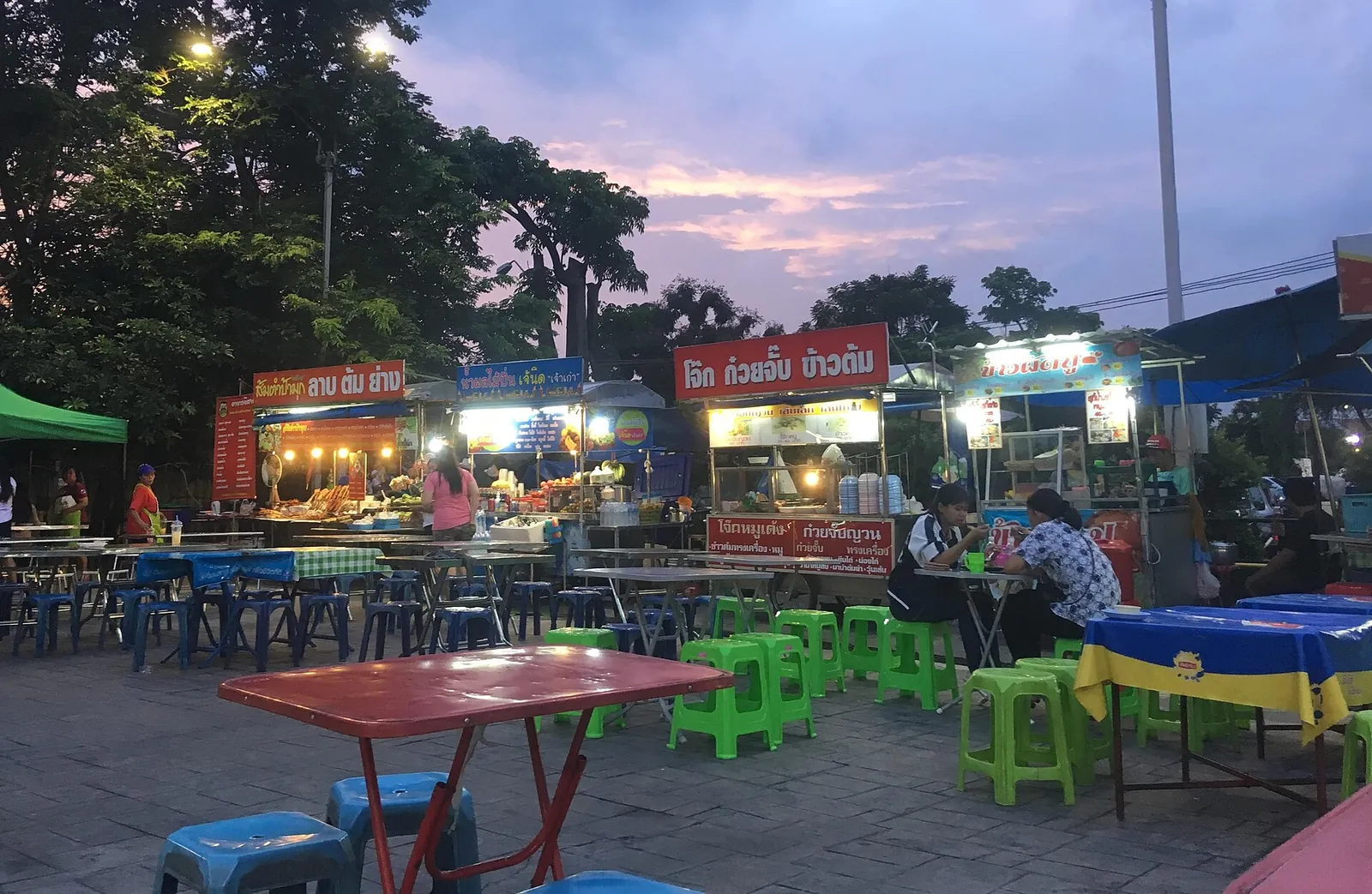 Food stalls with plastic tables and stools at an Ubon night market at dusk