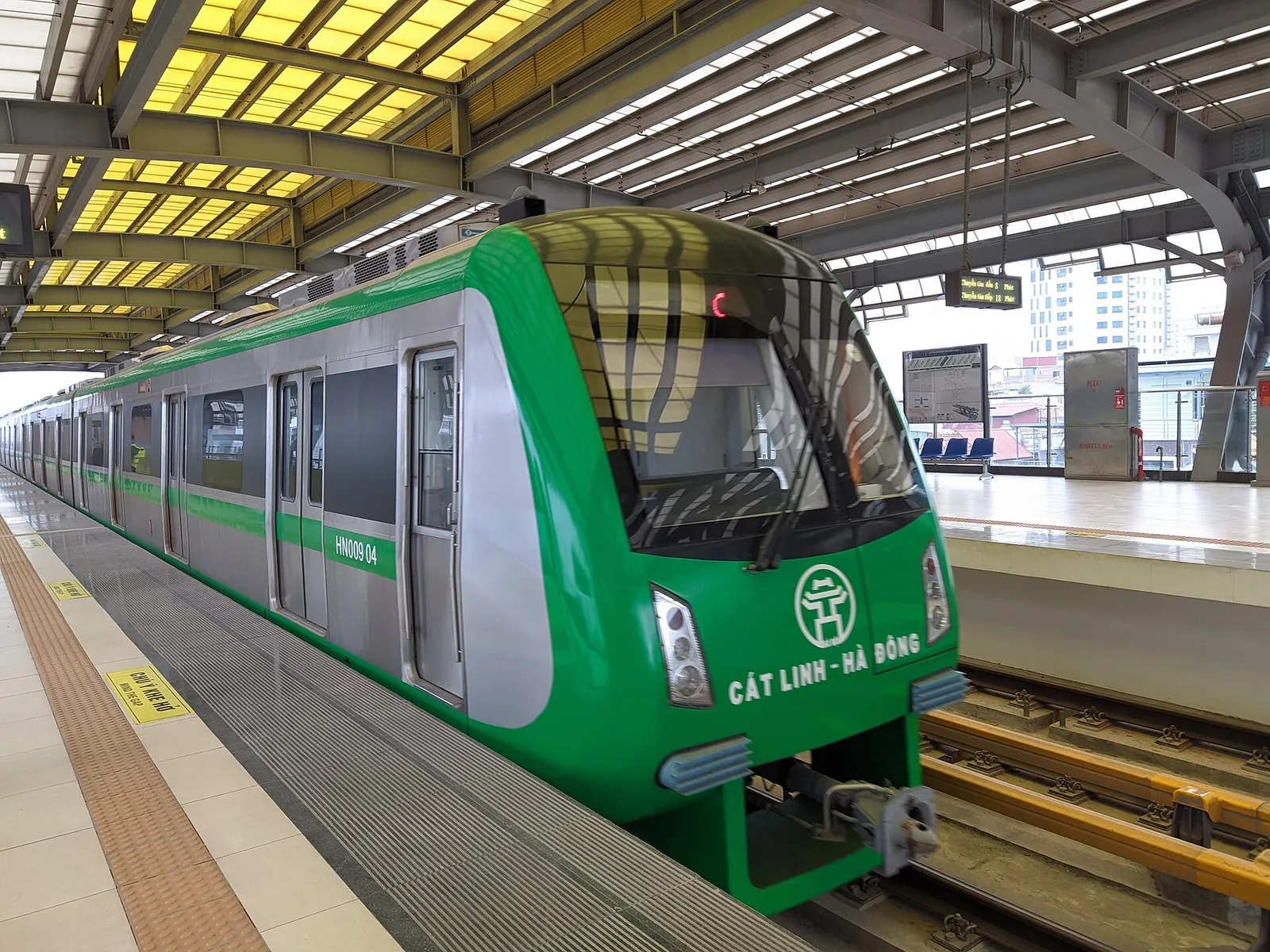 Green Hanoi Metro Line 2A train arriving at an elevated platform with a yellow tactile strip at the platform edge