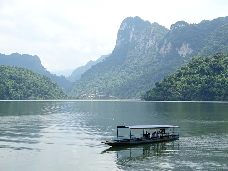 A small passenger boat crossing Ba Be Lake below forested limestone cliffs