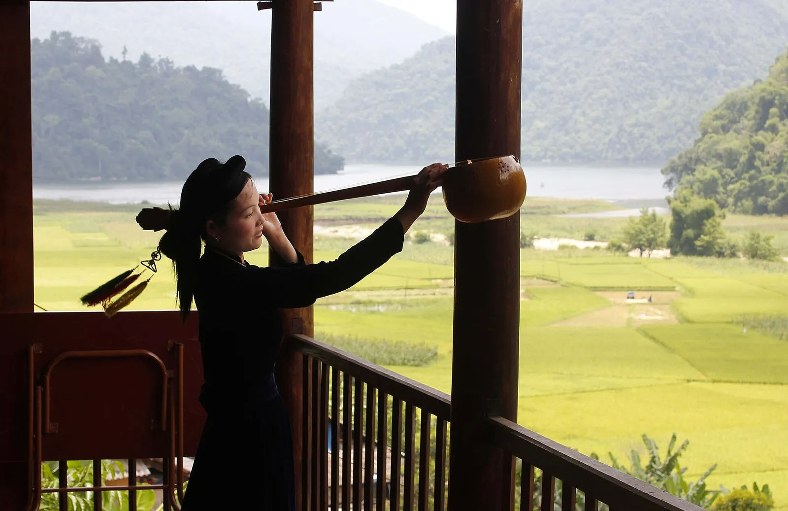 A Tay performer in dark dress playing a long-necked tinh lute on a wooden balcony above green fields and the lake