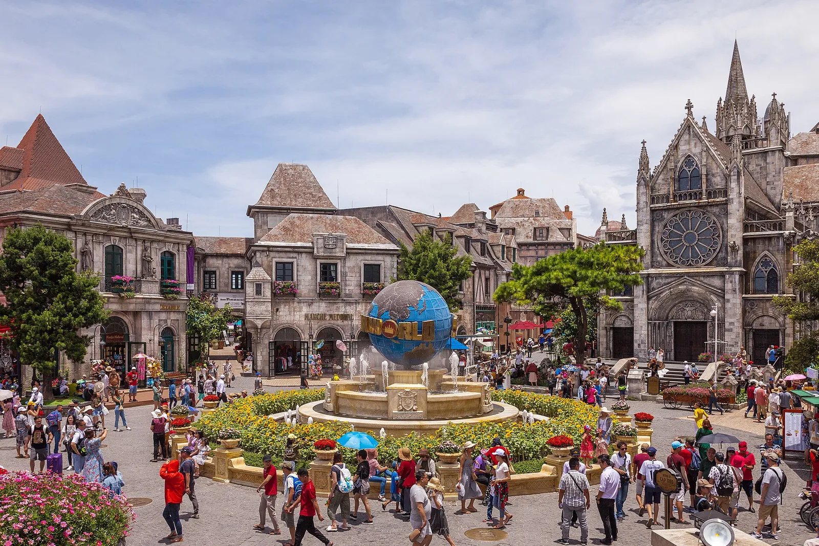 Crowds in the main square of the French Village at Ba Na Hills, with a globe fountain, flower beds and a Gothic-style church