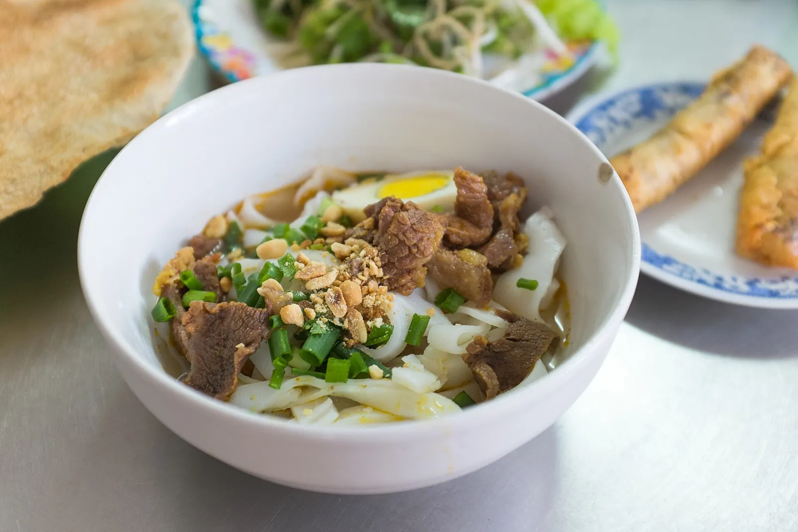 A white bowl of mi Quang in Da Nang with wide rice noodles, sliced meat, a quail egg, peanuts and spring onion, with a fried roll and a rice cracker beside it