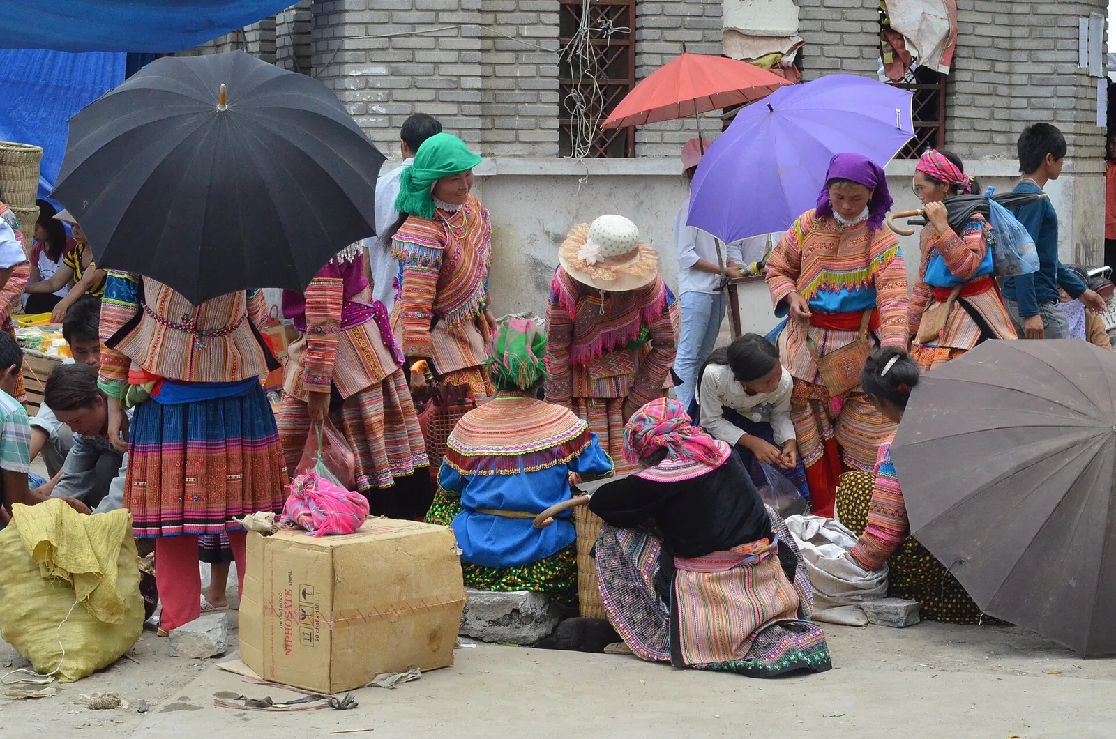 Flower Hmong women in bright embroidered skirts and headscarves selling goods under umbrellas at Bac Ha Sunday market